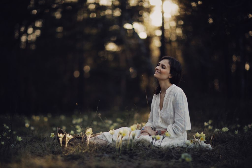A woman sits peacefully in a sunlit forest clearing surrounded by flowers.